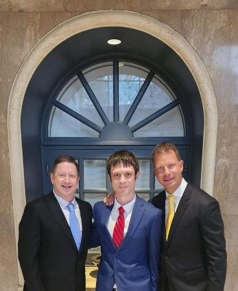 Three professionals posing together in formal attire, with one wearing a blue suit and red tie, standing in front of a decorative window, symbolizing partnership and collaboration in business strategy.